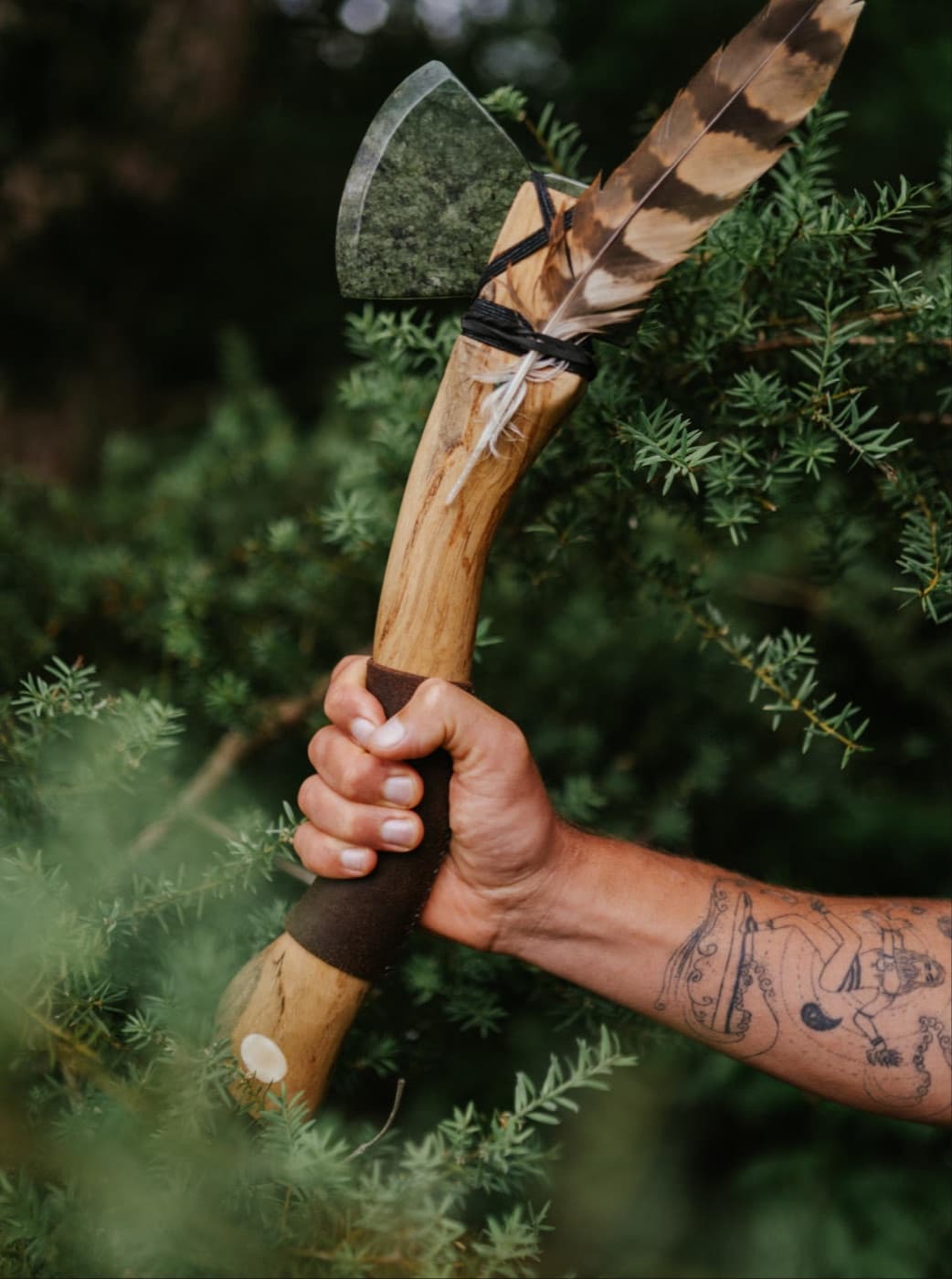 A hand holding a pounamu axe bound with a feather, against green bush